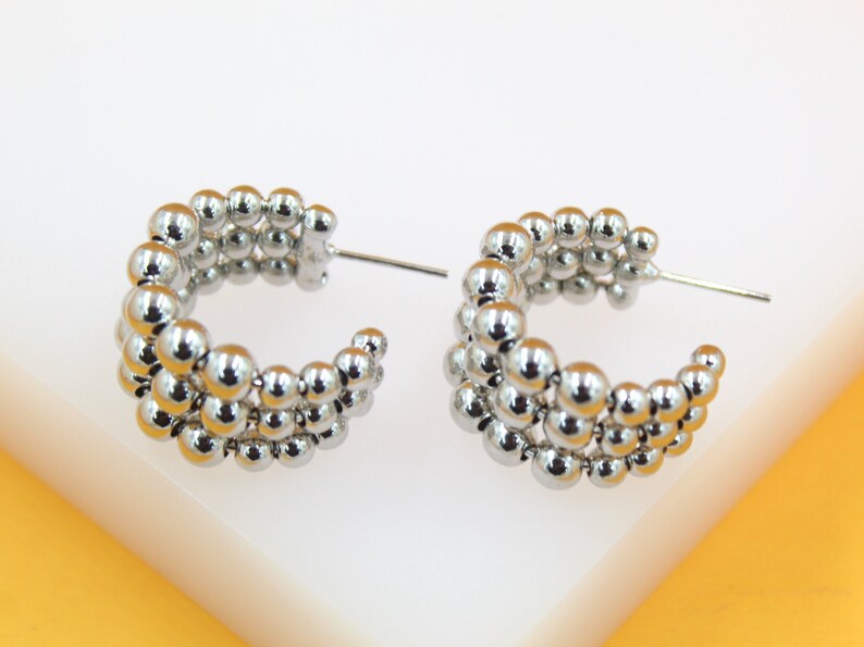 A pair of glossy silver beaded hoop earrings rest against a clean white and orange background.