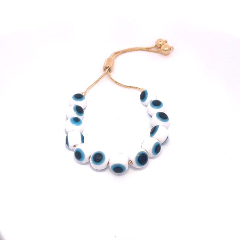 A delicate gold-toned adjustable slider bracelet features a string of white and blue glass evil eye beads against a plain white background.