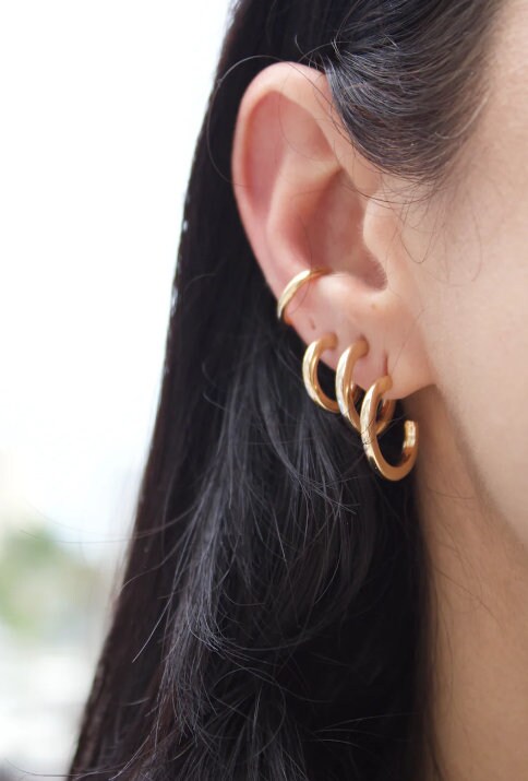 A person showcases a curated ear stack featuring stacked gold hoop earrings and a sleek upper ear cuff.