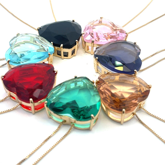 Seven vibrant heart-shaped crystal necklaces in a rainbow of colors radiate outwards on fine gold chains against a white background.