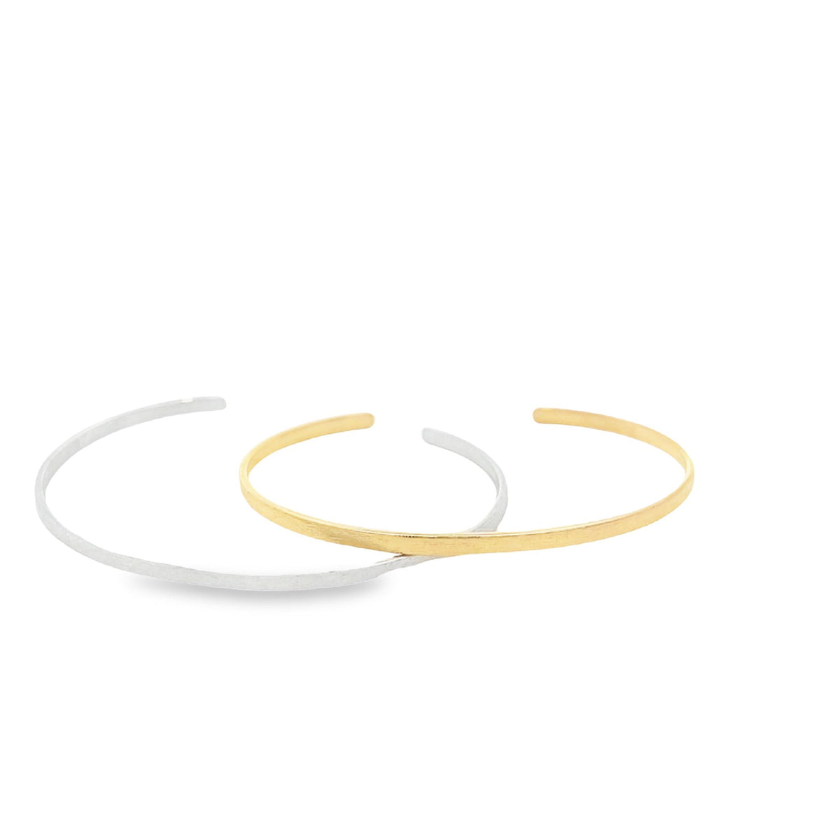 Two minimalist cuff bracelets, one in polished silver and the other in gold, rest overlapping on a clean white background.