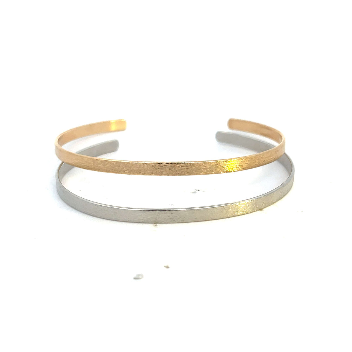 A gold cuff bracelet and a silver cuff bracelet rest stacked on a plain white surface.