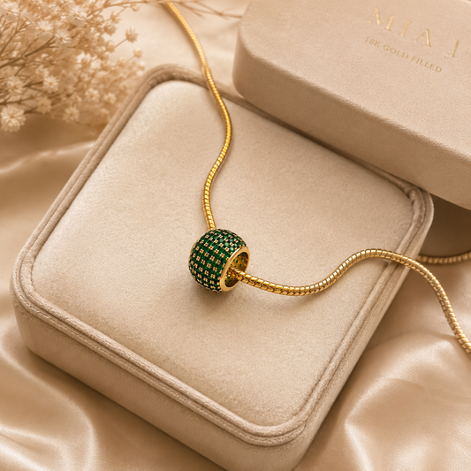 An 18k gold-filled Mia J necklace rests delicately on a beige velvet jewelry box, featuring a striking green crystal-embellished bead pendant.