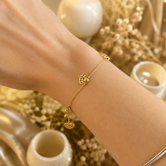 A delicate gold charm bracelet featuring intricate heart-shaped details dangles gracefully from a woman's wrist against a warm, neutral background.