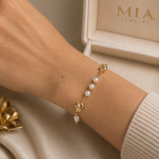 A delicate gold bracelet featuring pearls and teddy bear charms adorns a person's wrist near an open Mia Jewelry box.