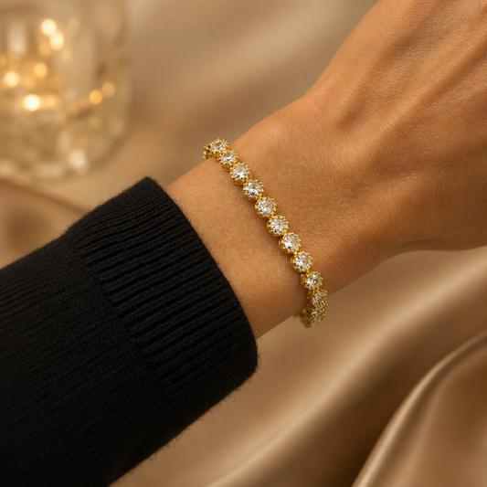 A woman wears a sparkling gold tennis bracelet adorned with round-cut cubic zirconia stones on her wrist against a warm satin background.