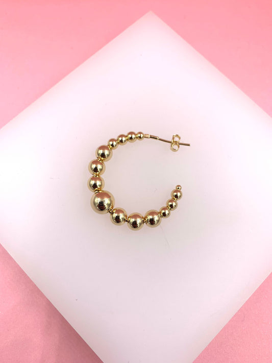 A single gold beaded hoop earring rests on a white display against a pink background.