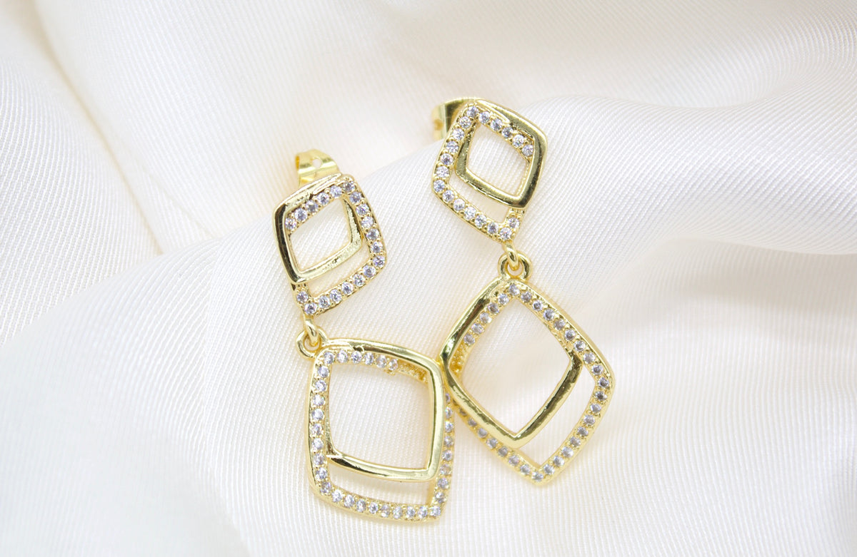 Two dangling gold diamond earrings rest on a soft white satin fabric background.