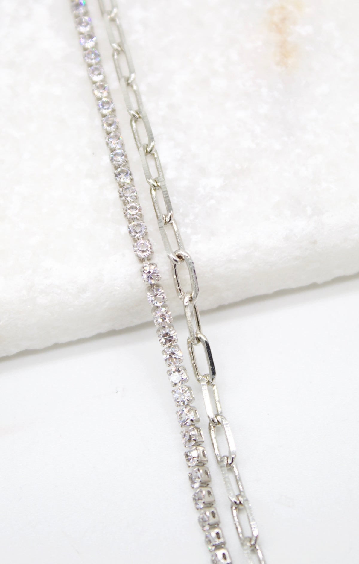 A silver tennis necklace layered with a paperclip chain rests against a white marble surface.