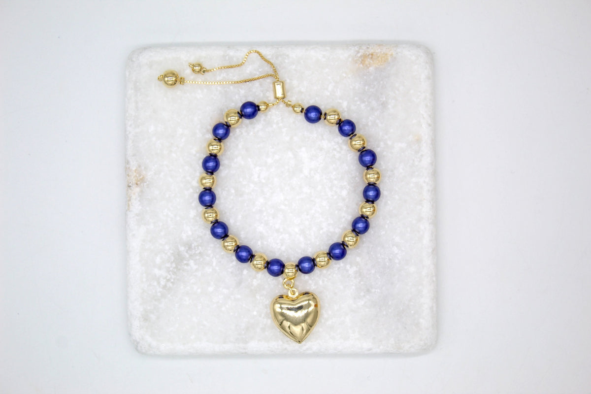 A handmade blue and gold beaded bolo bracelet with a dangling gold heart charm rests on a white marble slab.