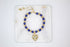 A handmade blue and gold beaded bolo bracelet with a dangling gold heart charm rests on a white marble slab.