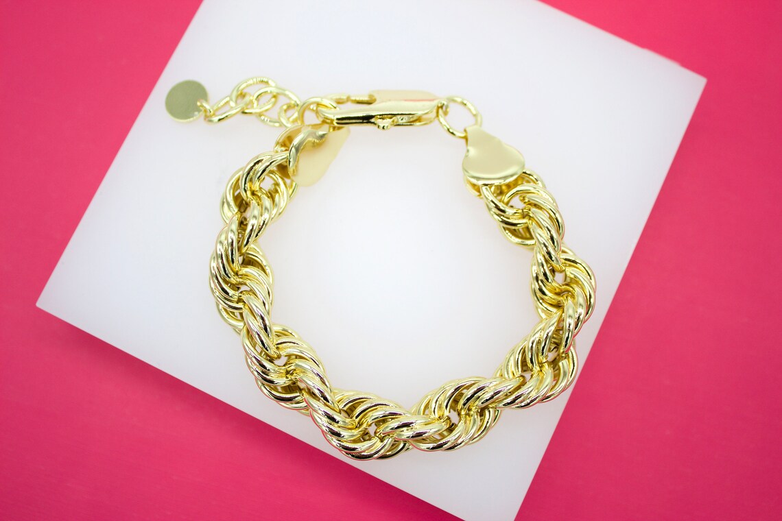 A thick, gold-plated rope chain bracelet rests on a white display against a vibrant pink background.