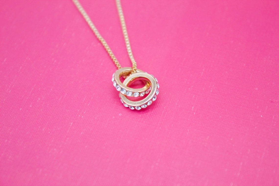 A delicate gold chain holds an interlocking double-ring pendant embellished with sparkling crystals against a vibrant pink background.