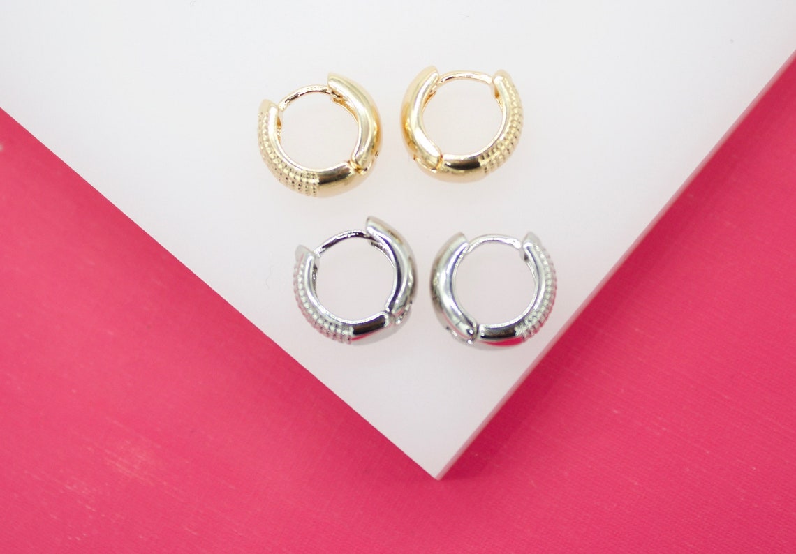 Two pairs of small huggie hoop earrings, one gold and one silver with textured detailing, rest on a white surface against a bright pink background.