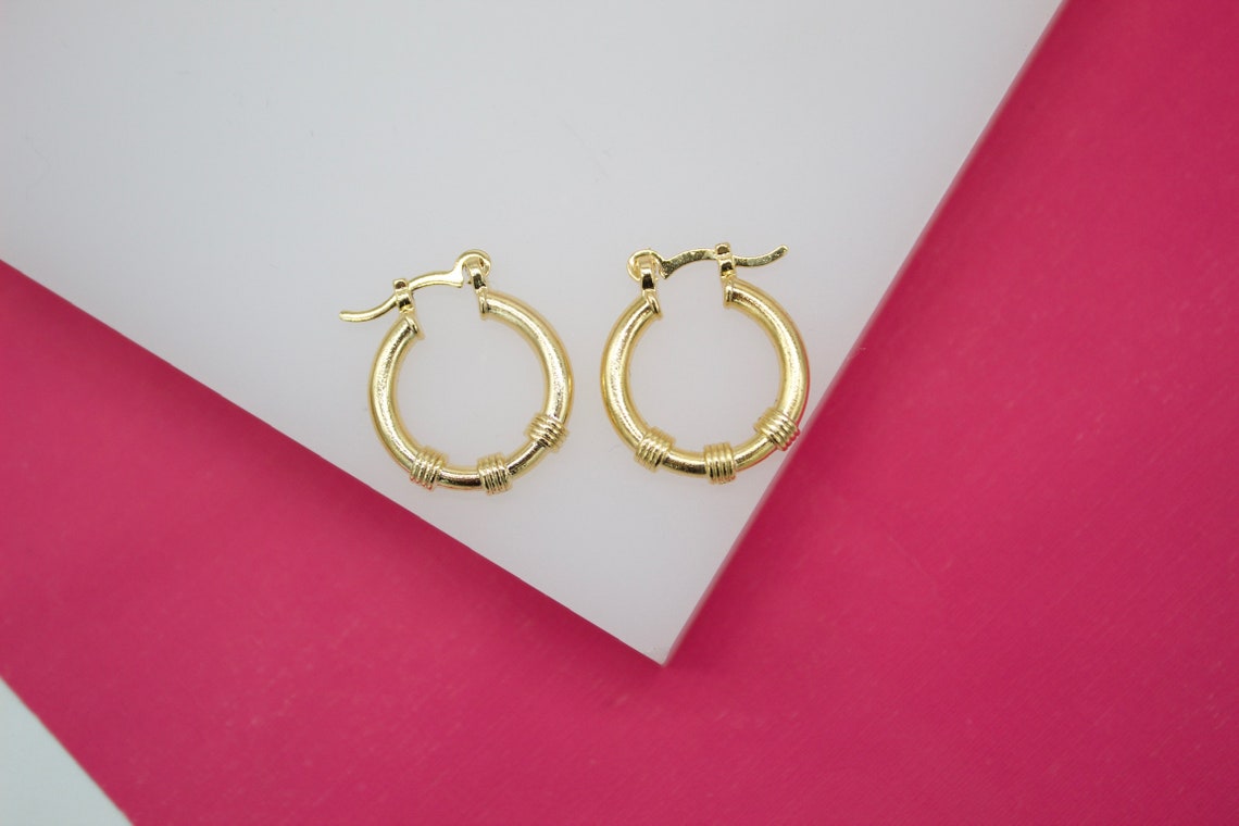 A pair of polished gold hoop earrings featuring decorative banded details rests on a white display surface against a vibrant pink background.