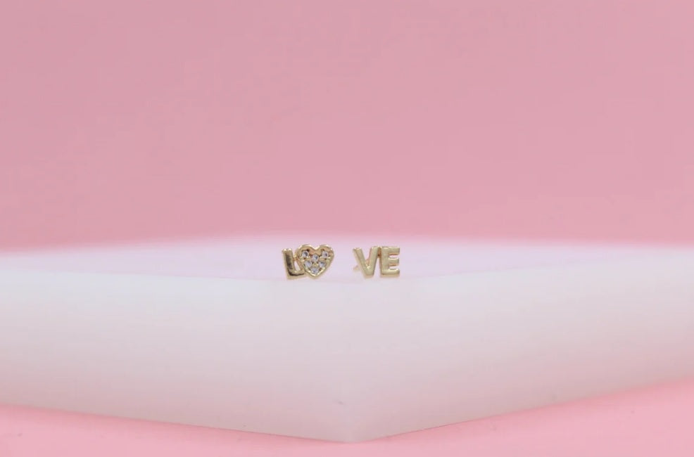 Gold stud earrings spelling "LOVE" with a pave diamond heart resting on a white surface against a pink background.