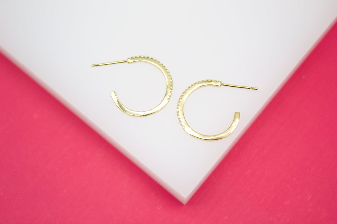 Two gold pave hoop earrings rest on a white surface against a bright pink background.