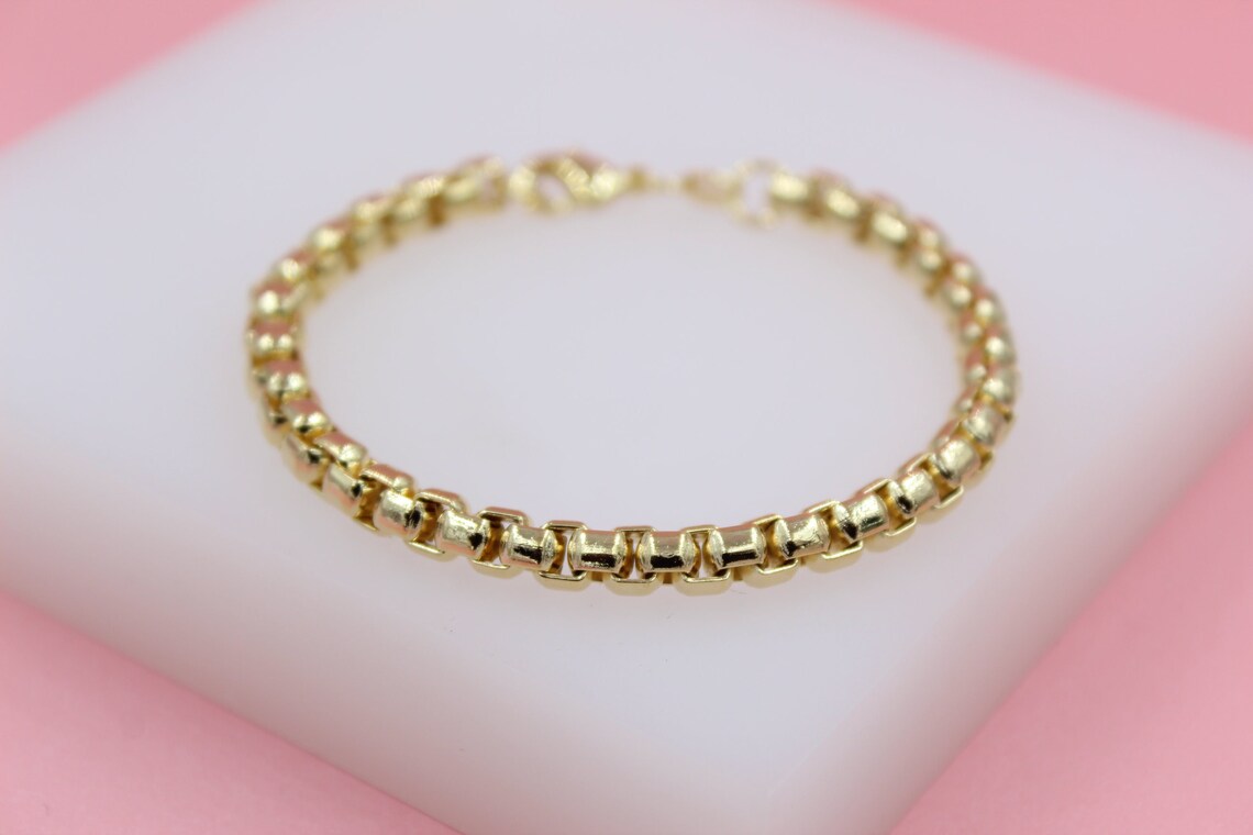 A polished gold box chain bracelet rests on a white display block against a soft pink background.