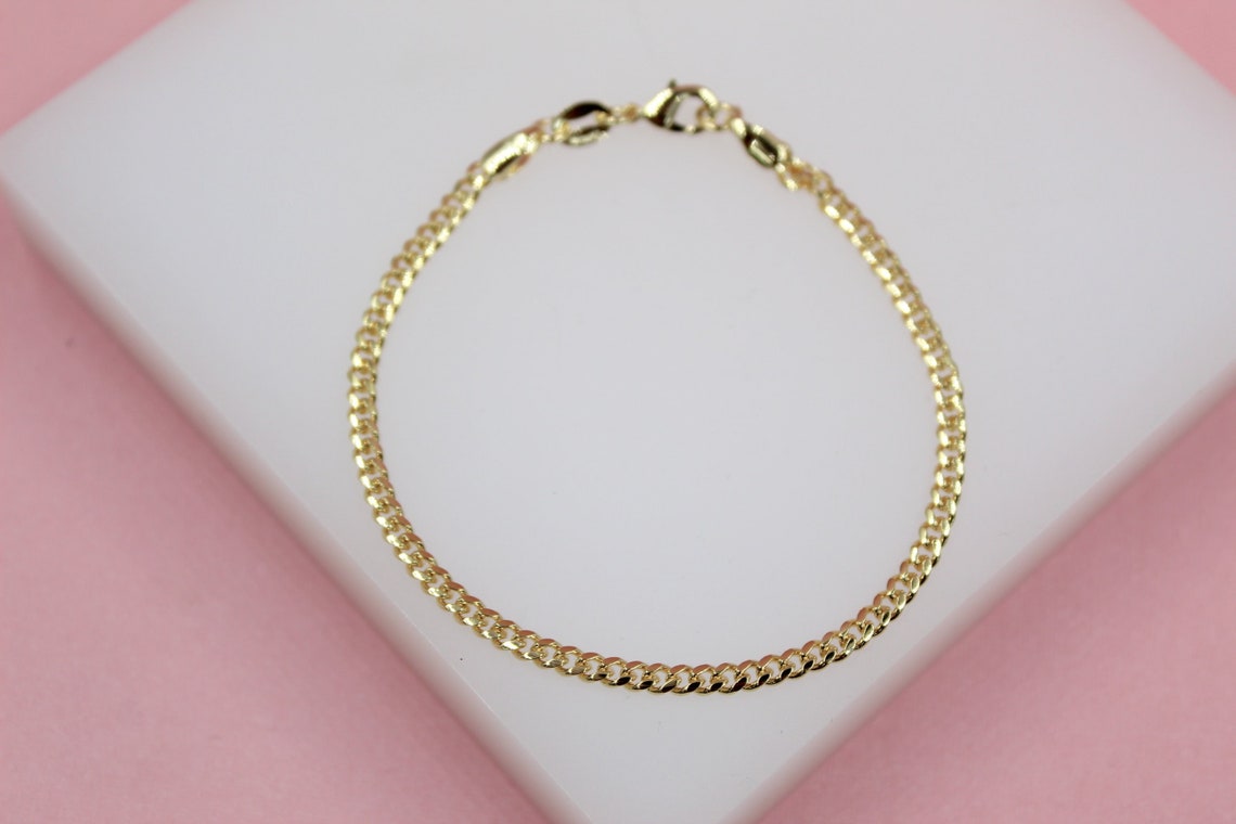 A delicate gold curb chain bracelet rests on a white display block against a soft pink background.