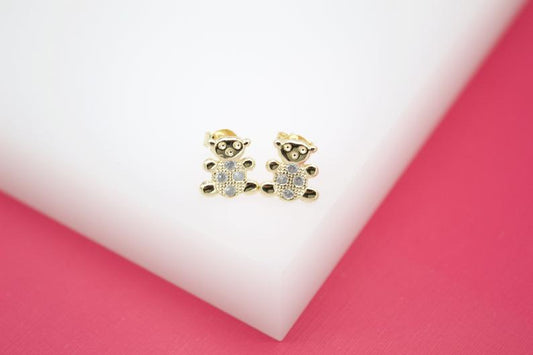 A pair of gold teddy bear earrings featuring sparkling cubic zirconia stones rests on a white display against a pink background.