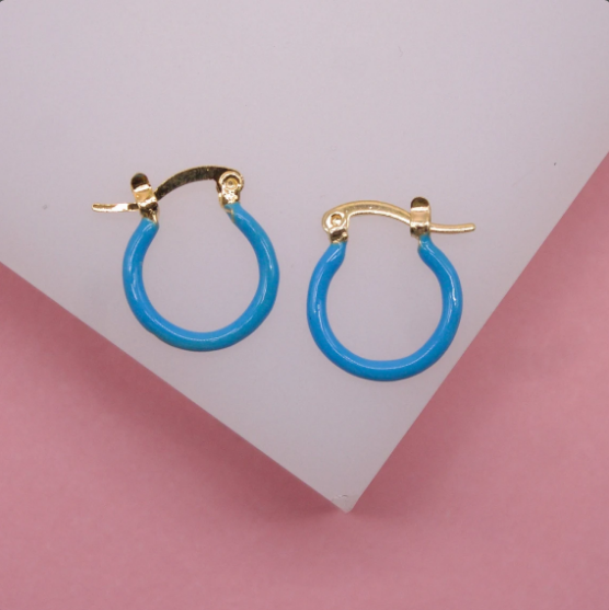 A pair of gold huggie hoop earrings featuring vibrant turquoise enamel inlay rest on a white surface against a pink background.