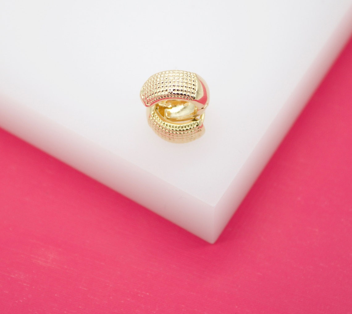 A gold textured huggie hoop earring rests on a white display block against a vibrant pink background.