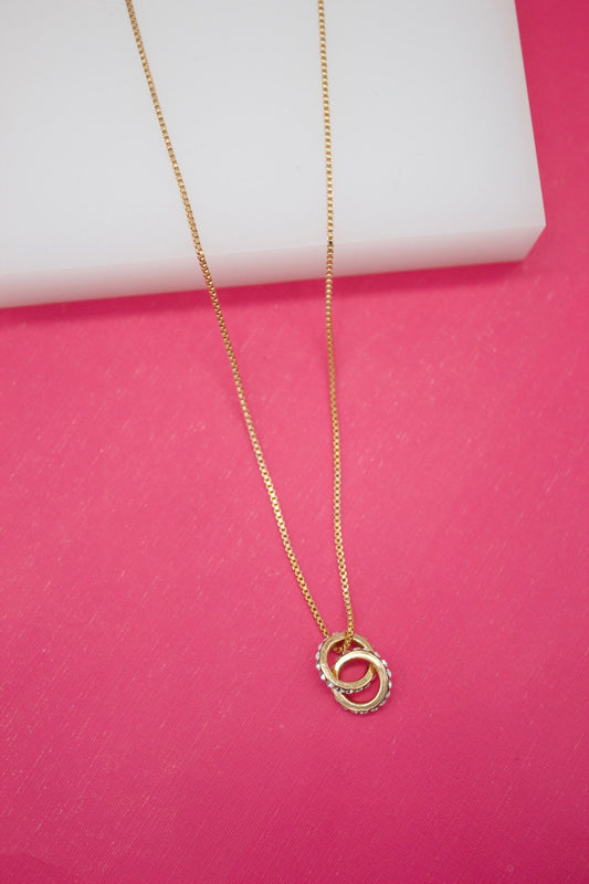 A delicate gold chain necklace with interlocking double-ring pendant rests against a bright pink background.