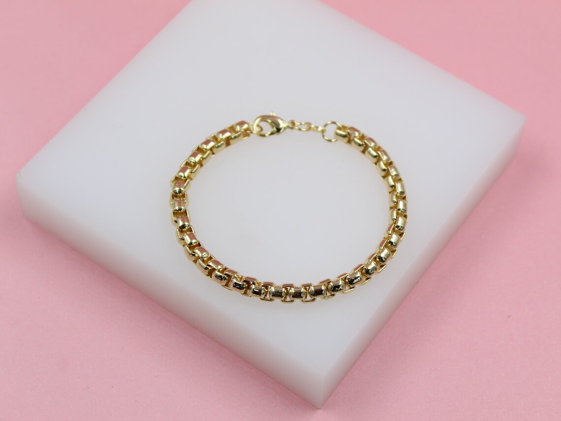 A polished gold box chain bracelet rests on a white block against a pink background.