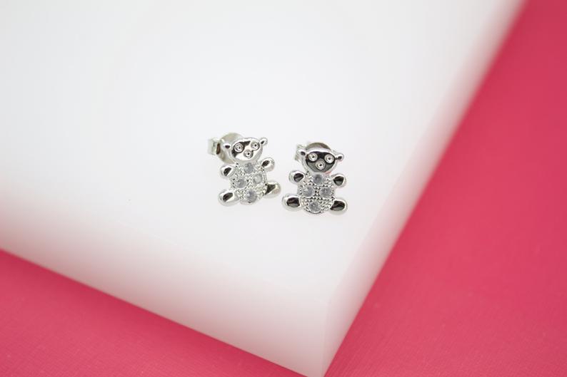 A pair of silver teddy bear stud earrings with cubic zirconia stones rest on a white display against a pink background.