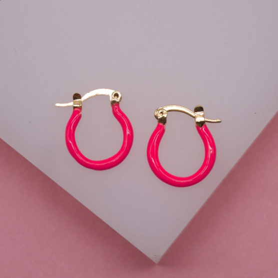 A pair of vibrant neon pink enamel and gold hoop earrings rest on a white surface against a pink background.
