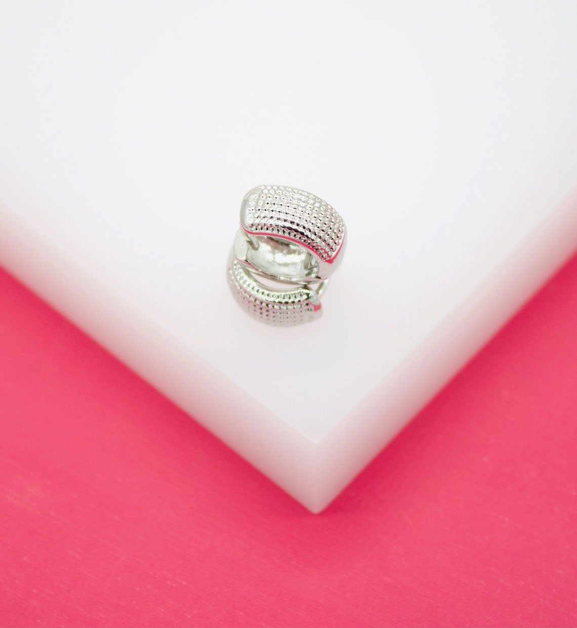 A textured silver huggie hoop earring rests on a white display block against a vibrant pink background.