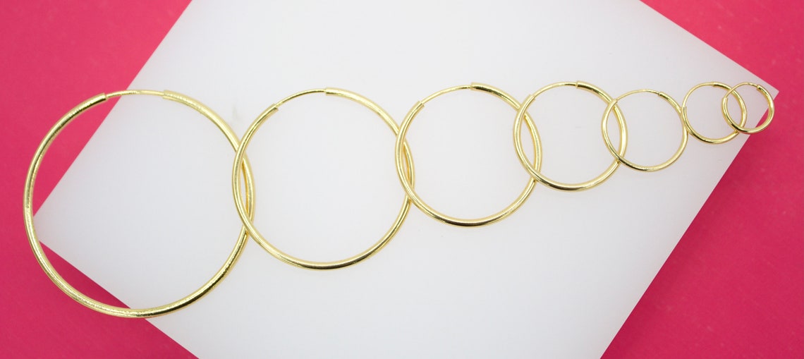 A graduated set of polished gold hoop earrings in descending sizes rests on a white display surface against a pink background.