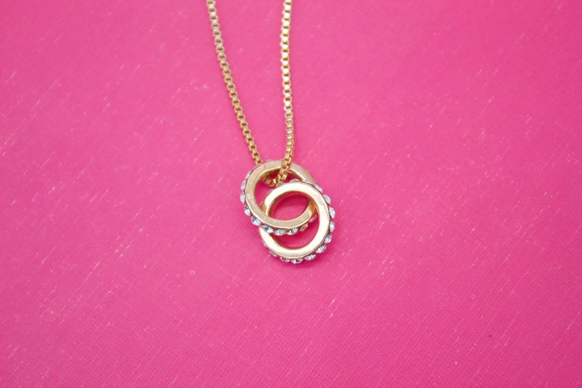 A delicate gold chain holds an interlocking double-ring pendant embellished with sparkling crystals against a vibrant pink background.