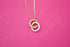 A delicate gold chain holds an interlocking double-ring pendant embellished with sparkling crystals against a vibrant pink background.