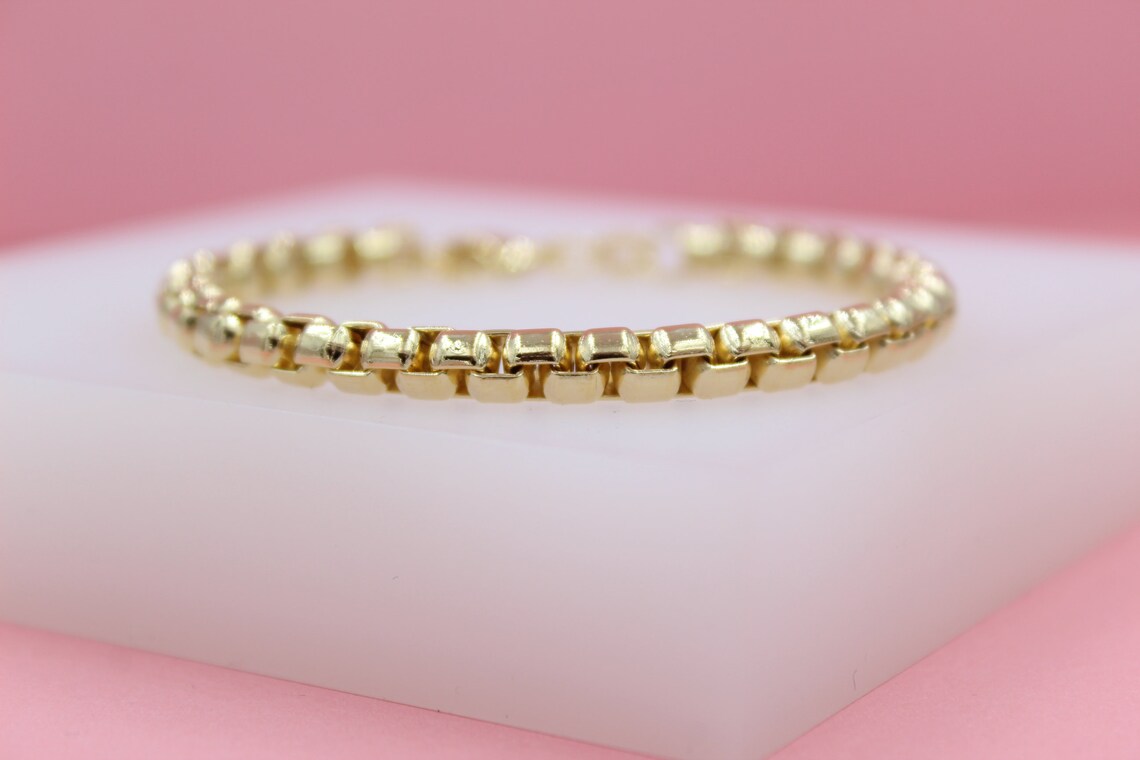 A polished gold Venetian box chain bracelet rests on a white display block against a soft pink background.