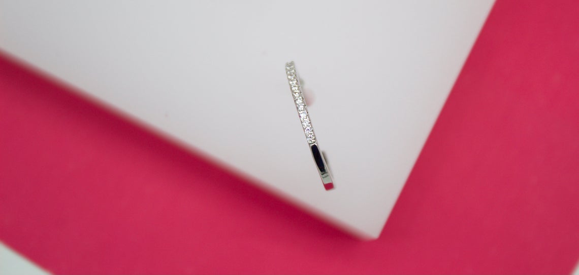 A delicate silver hoop earring encrusted with sparkling cubic zirconia stones rests on a white display against a vibrant pink background.