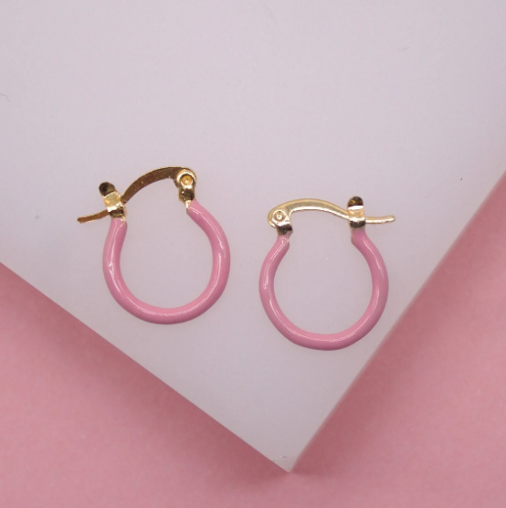 A pair of small gold hoop earrings with light pink enamel coating rests on a white and pink surface.