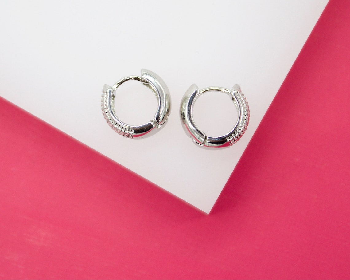 A pair of small silver hoop earrings rests on a white display block against a vibrant pink background.