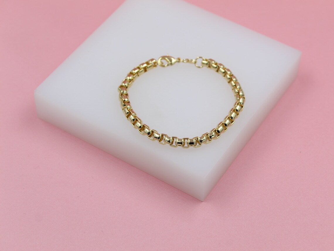 A delicate gold chain bracelet rests on a white display block against a soft pink background.