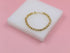A delicate gold chain bracelet rests on a white display block against a soft pink background.
