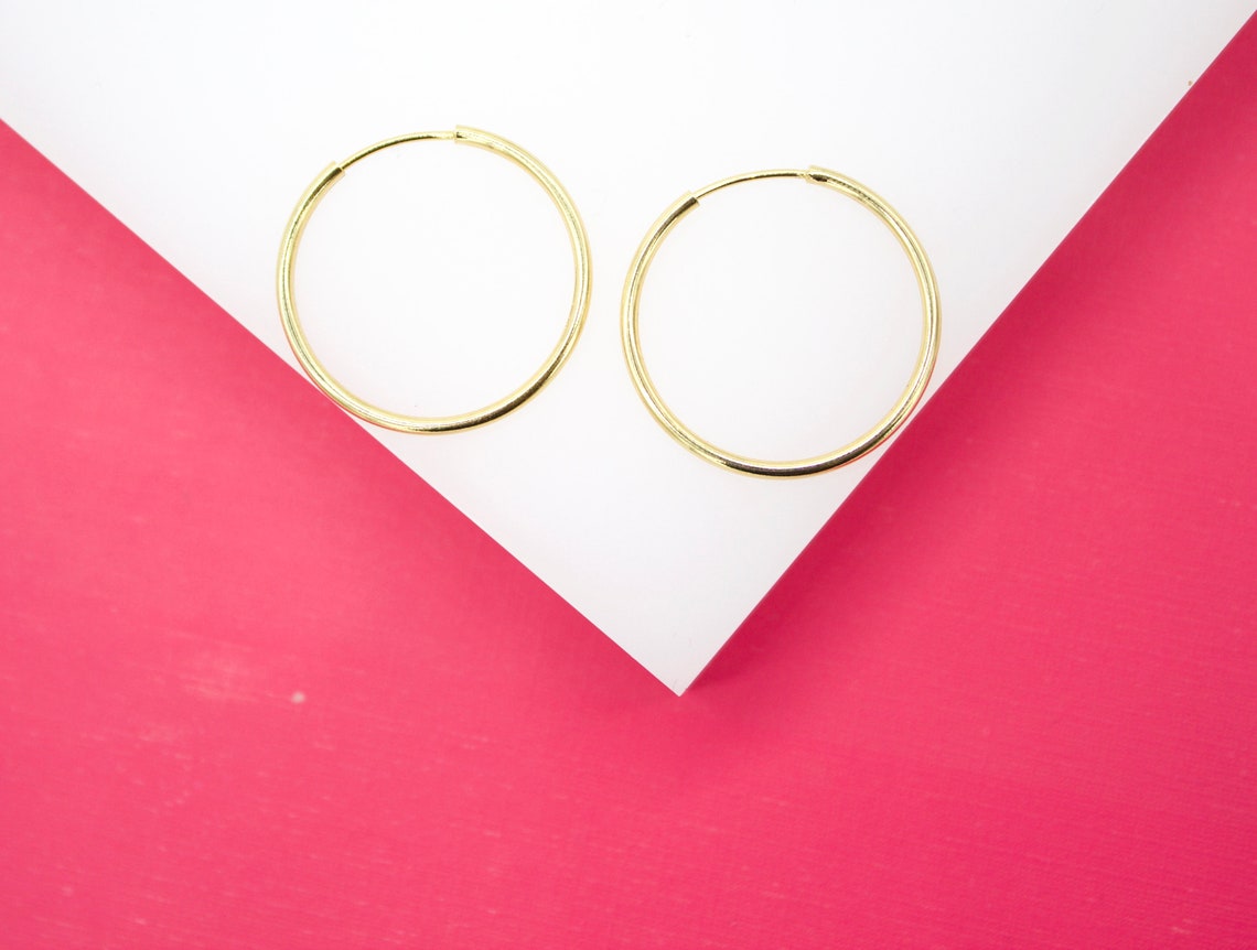 Two minimalist gold hoop earrings rest against a crisp white and vibrant pink geometric backdrop.