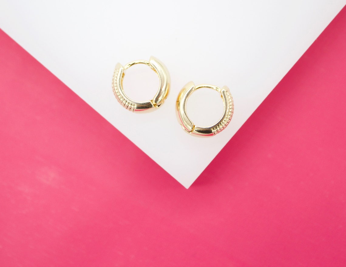 A pair of small gold huggie hoop earrings with textured detail rests on a white and pink geometric display surface.