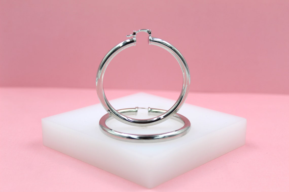 Two polished silver hoop earrings rest on a white display block against a soft pink background.