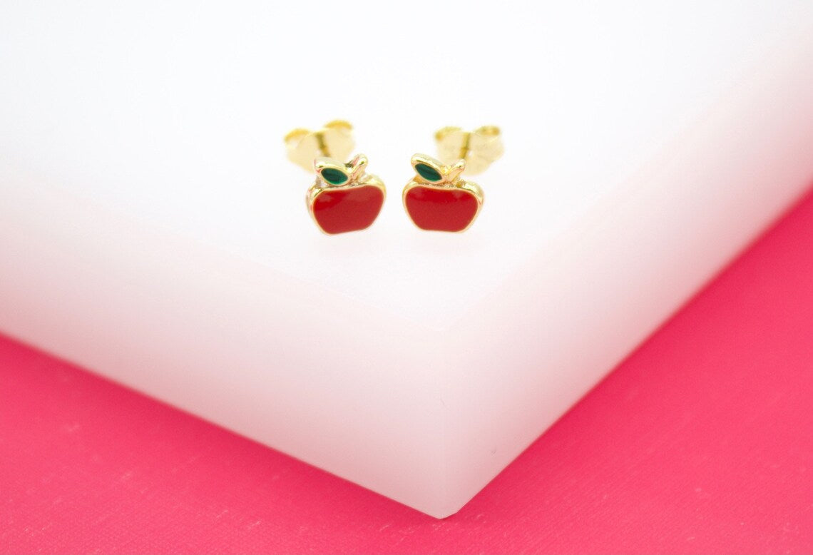 A pair of gold apple stud earrings with red enamel bodies and green leaves rests on a white and pink display stand.