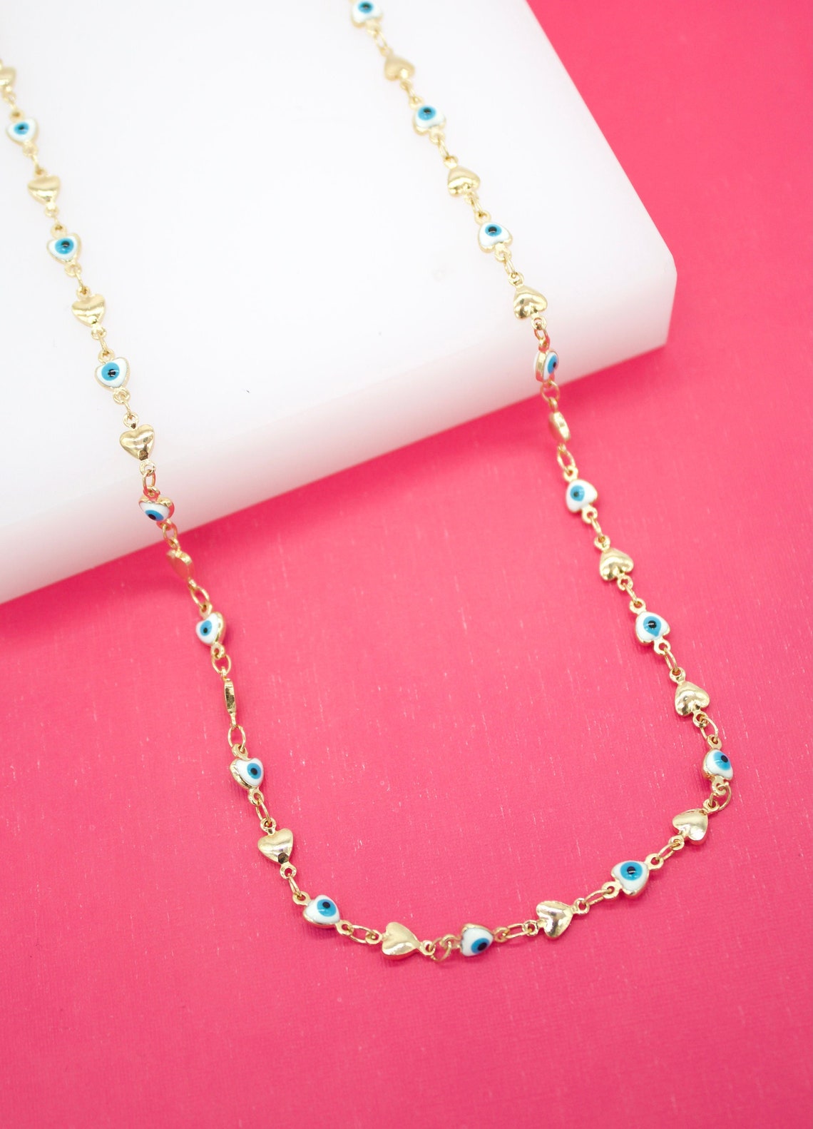 A gold chain necklace displays alternating evil eye charms and tiny heart links against a vibrant pink and white background.