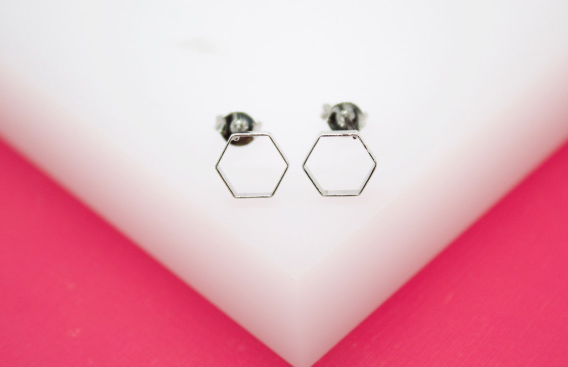 A pair of minimalist silver geometric hexagon stud earrings rests on a white and pink display surface.