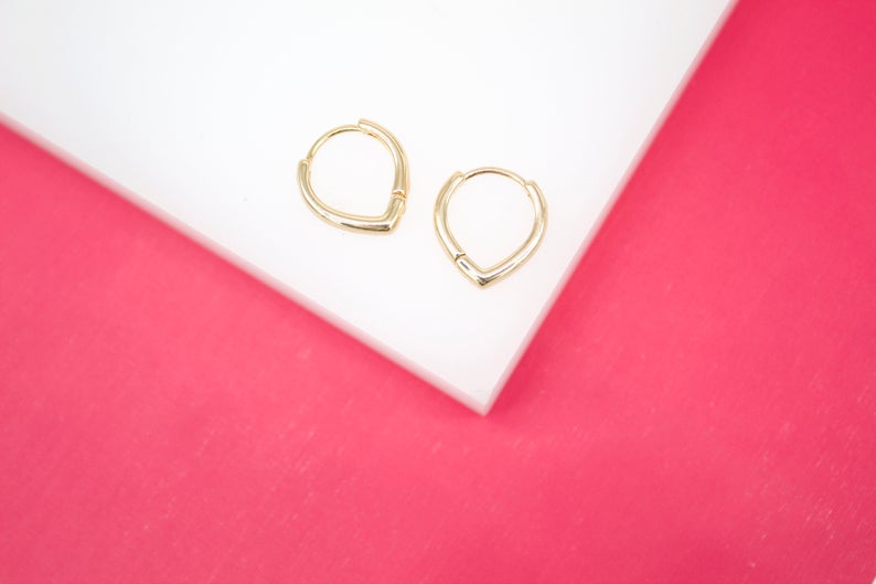 A pair of gold V-shaped hoop earrings rests on a white display surface against a vibrant pink background.
