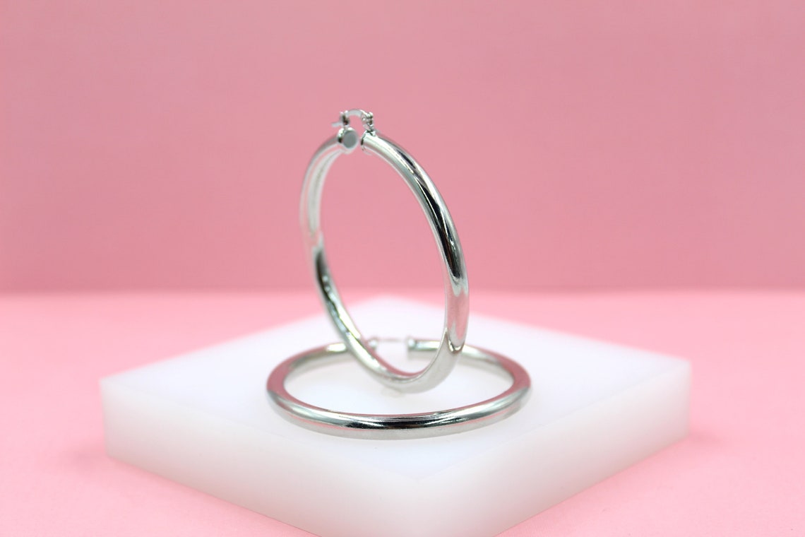 Two polished silver hoop earrings rest on a white display block against a soft pink background.