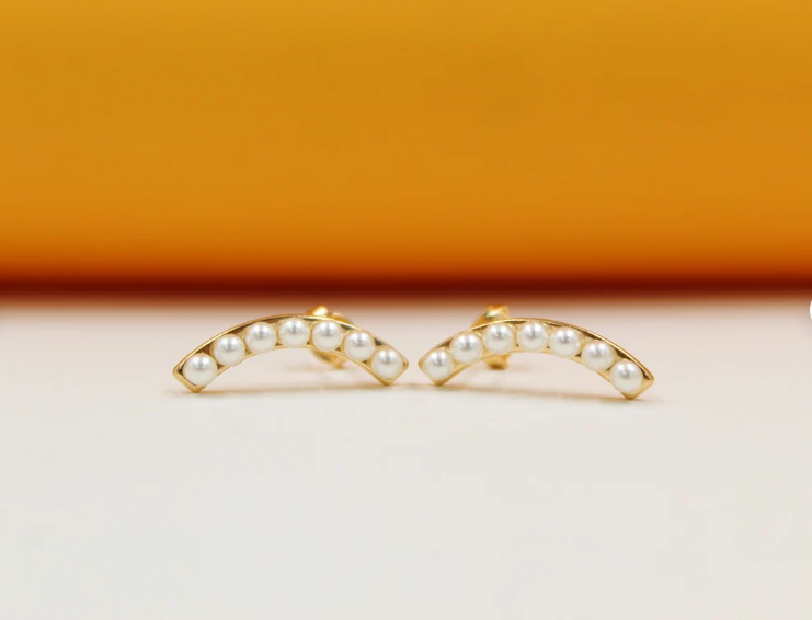 A pair of curved gold stud earrings featuring a row of small white pearls rests against a warm peach-toned background.