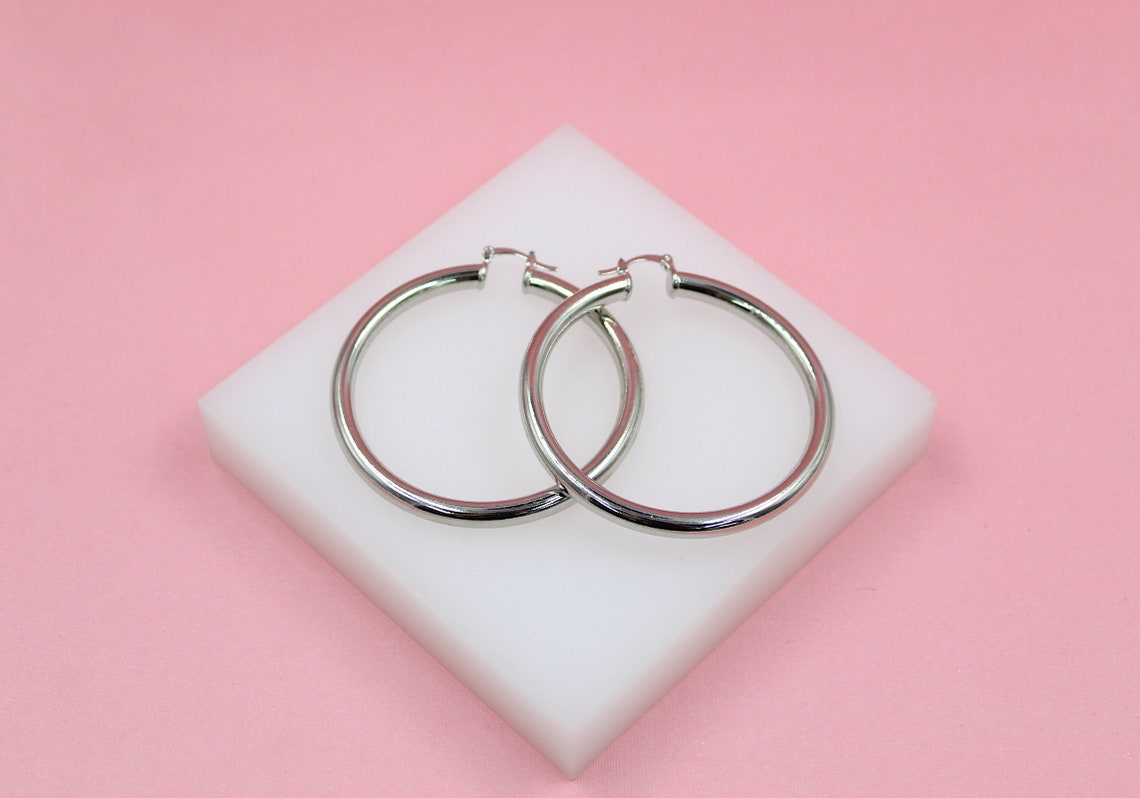 A pair of shiny silver hoop earrings rests on a white square display block against a soft pink background.